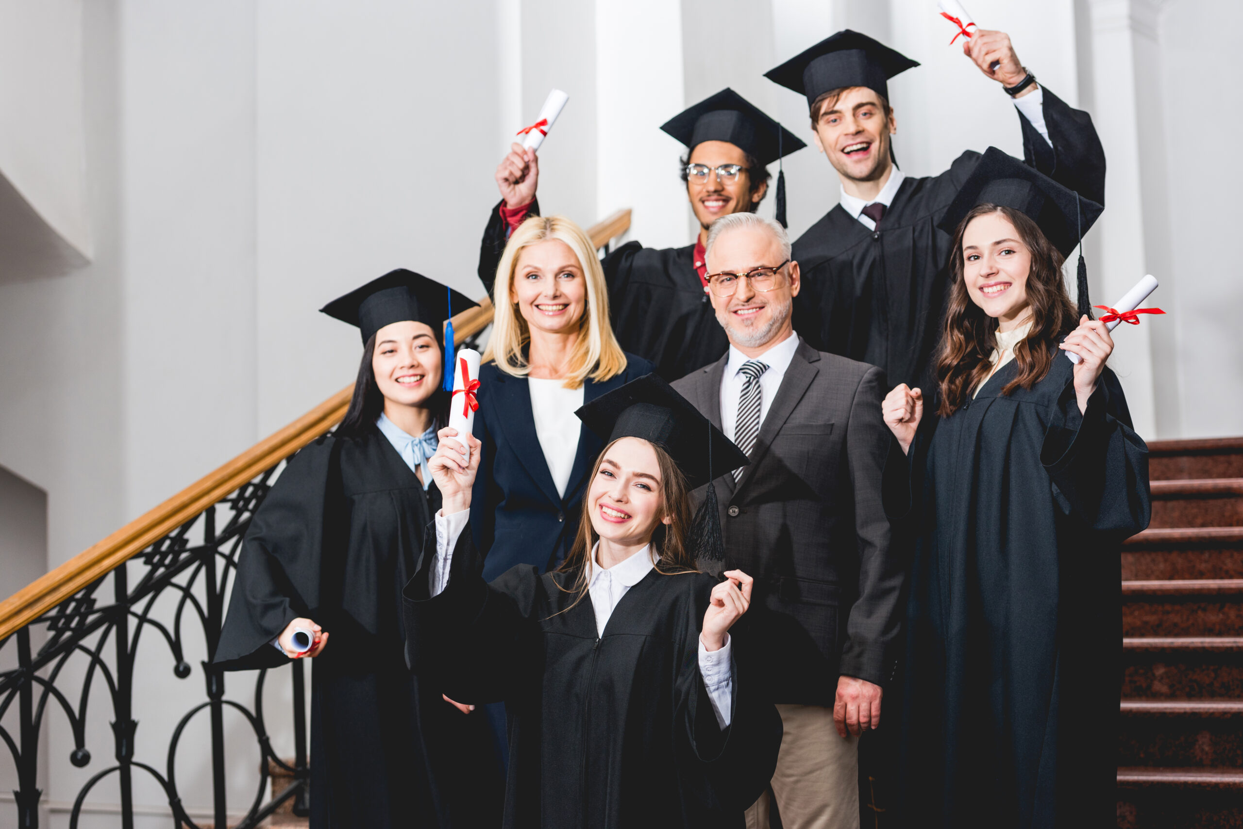 cheerful students in graduation caps holding diplomas near happy teachers