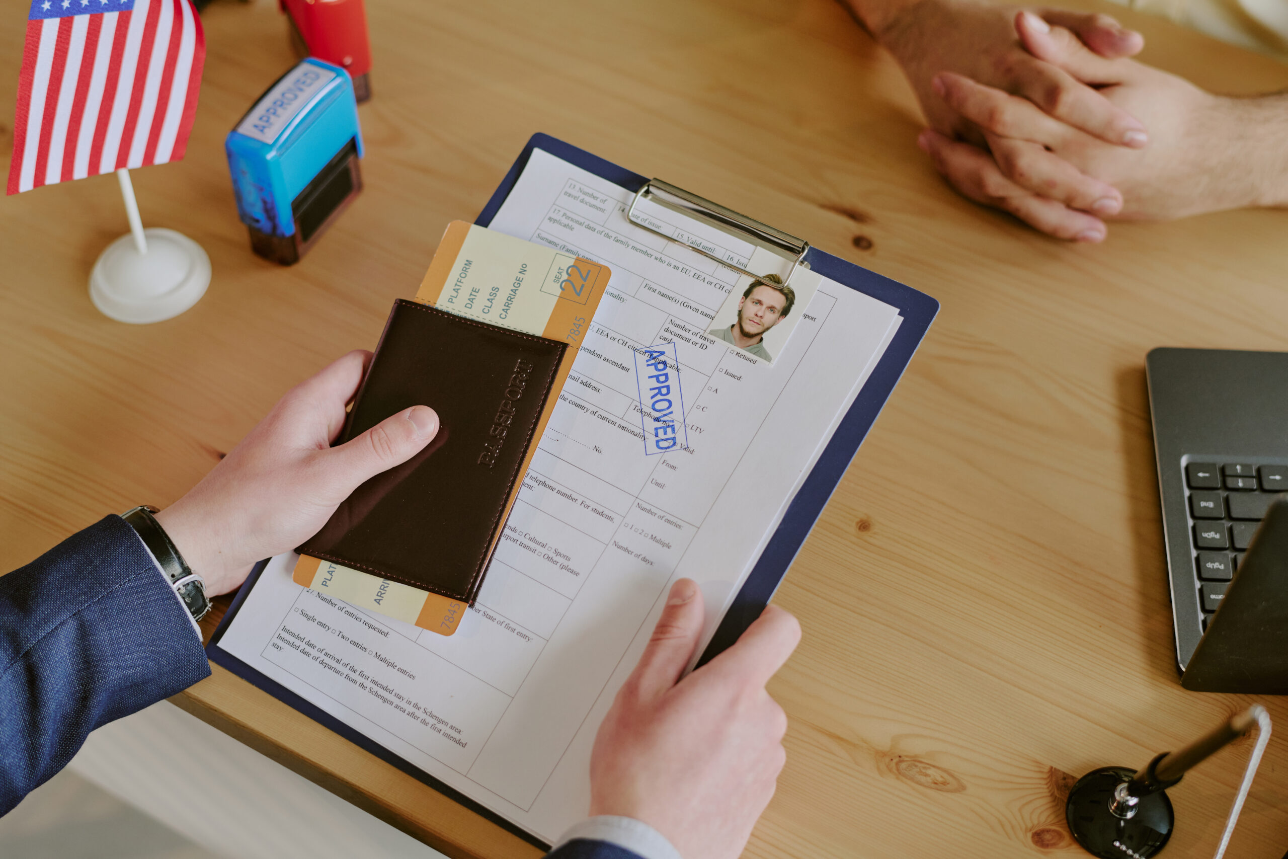Handing passport and documents across table during visa interview. American flag on table alongside laptop, envelope, and clipboard with approved form visible