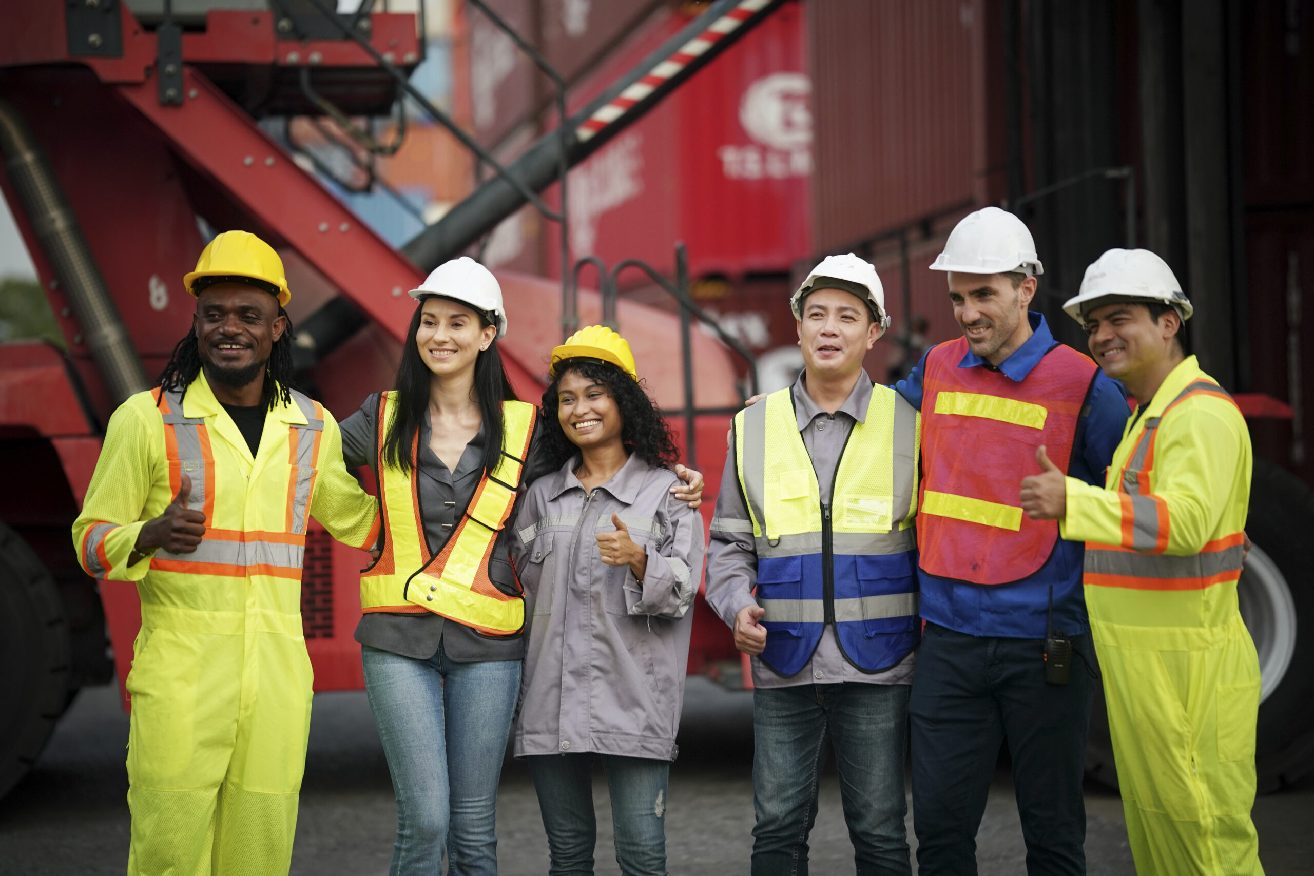 A group of workers proudly pose together in a shipping yard, smiling and giving thumbs up.
